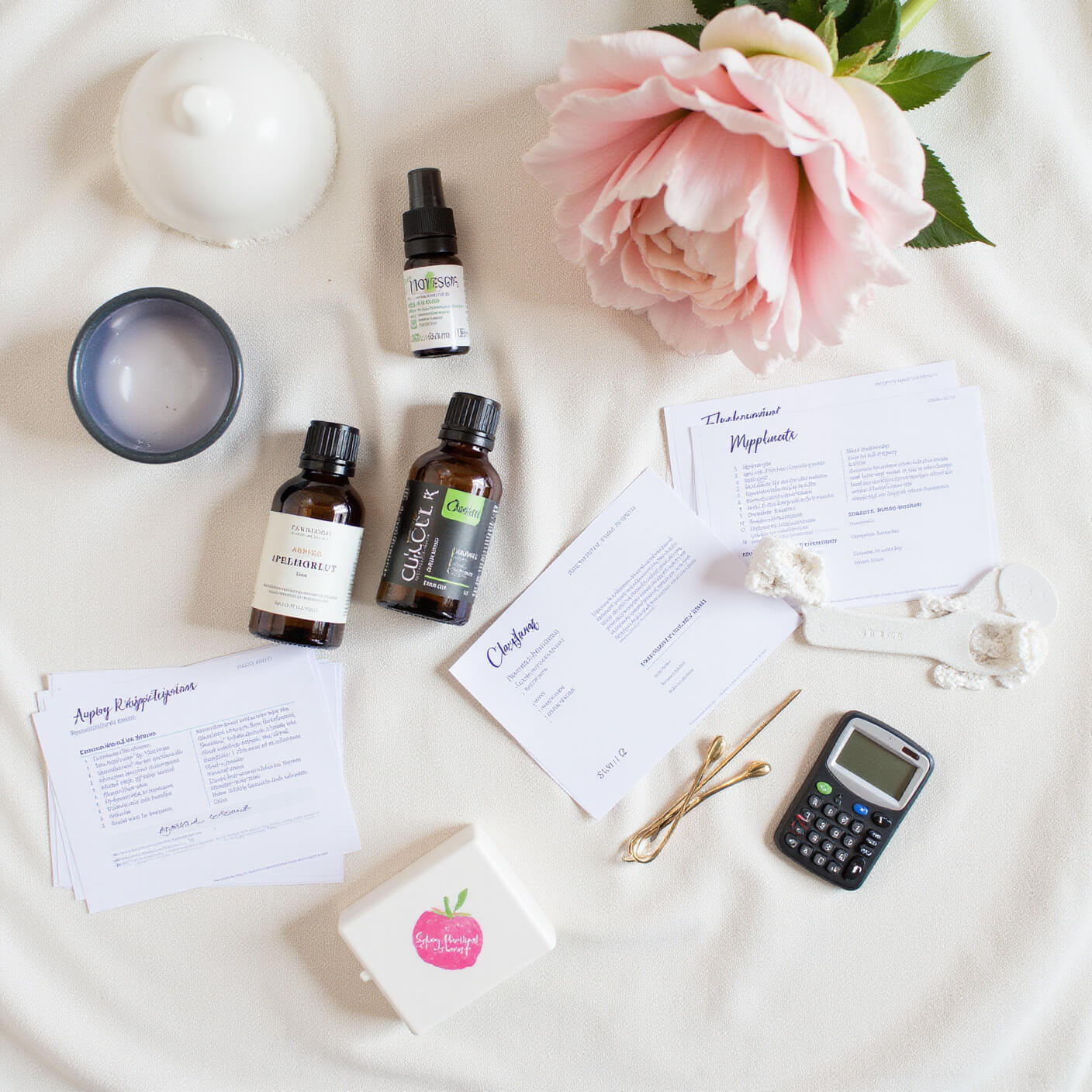 Overhead view of budget beauty supplies on linen: oils in simple bottles, DIY tools, mixing bowls, measuring spoons, a calculator, price tags, recipe cards, and a progress journal arranged neatly in bright light.
