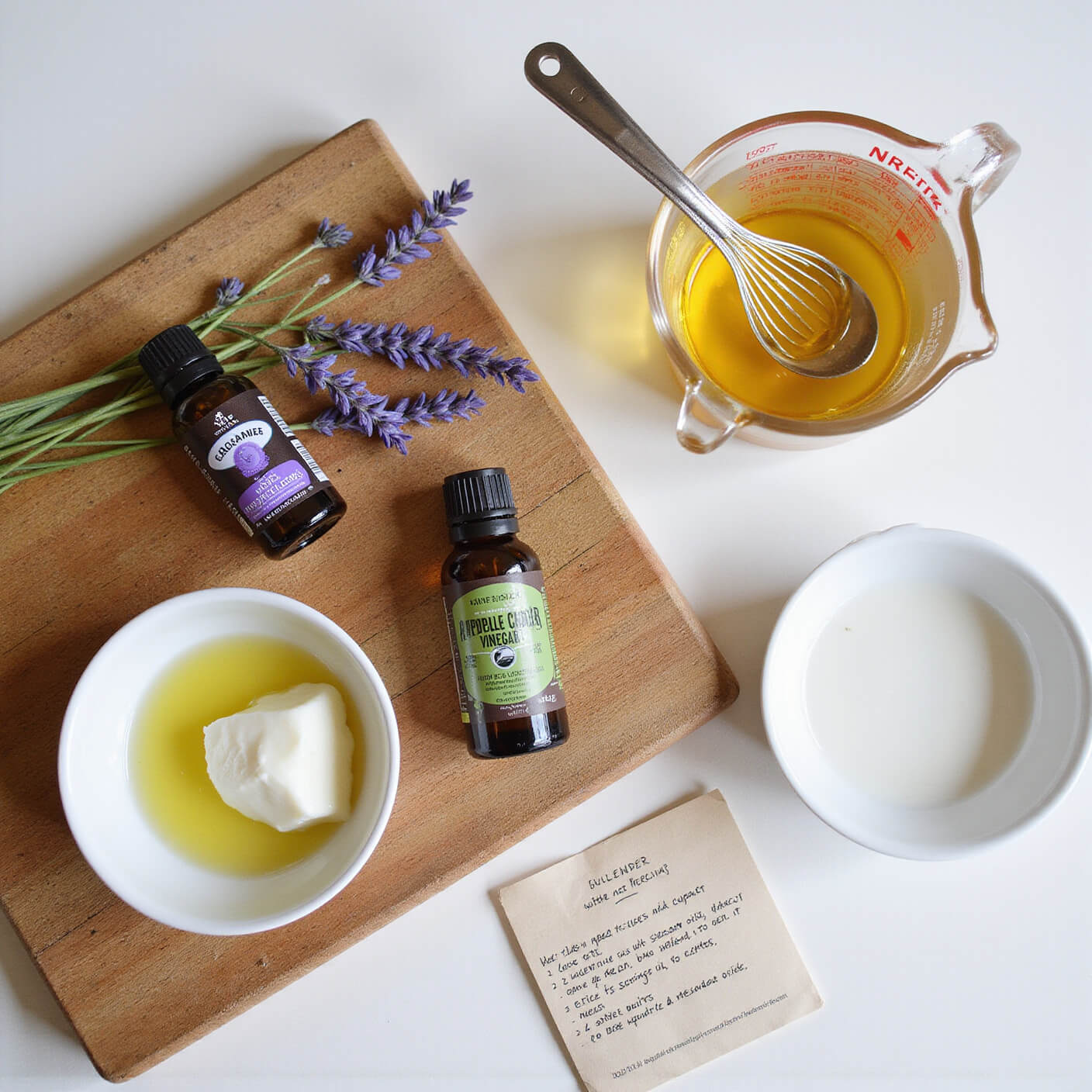 Overhead view of a modern farmhouse kitchen with essential oils, a measuring cup of vinegar and water, lavender sprigs, coconut and olive oils in white bowls, a handwritten recipe card, and a nit comb on a rustic cutting board.