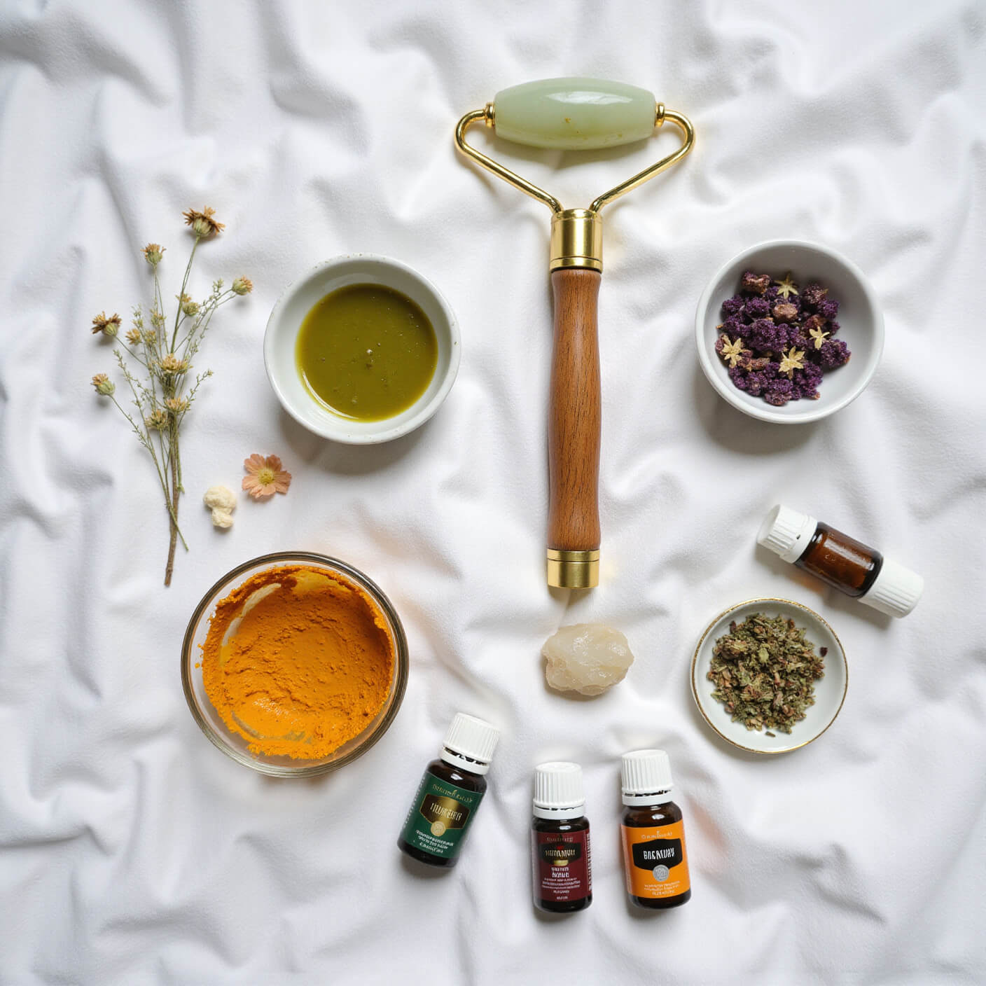 Overhead view of holistic skincare items arranged in a circle on rumpled white linen, featuring a wooden face brush, jade roller, bowls of turmeric and green tea masks, crystal clusters, dried flowers, and essential oil bottles, softly lit with natural light.