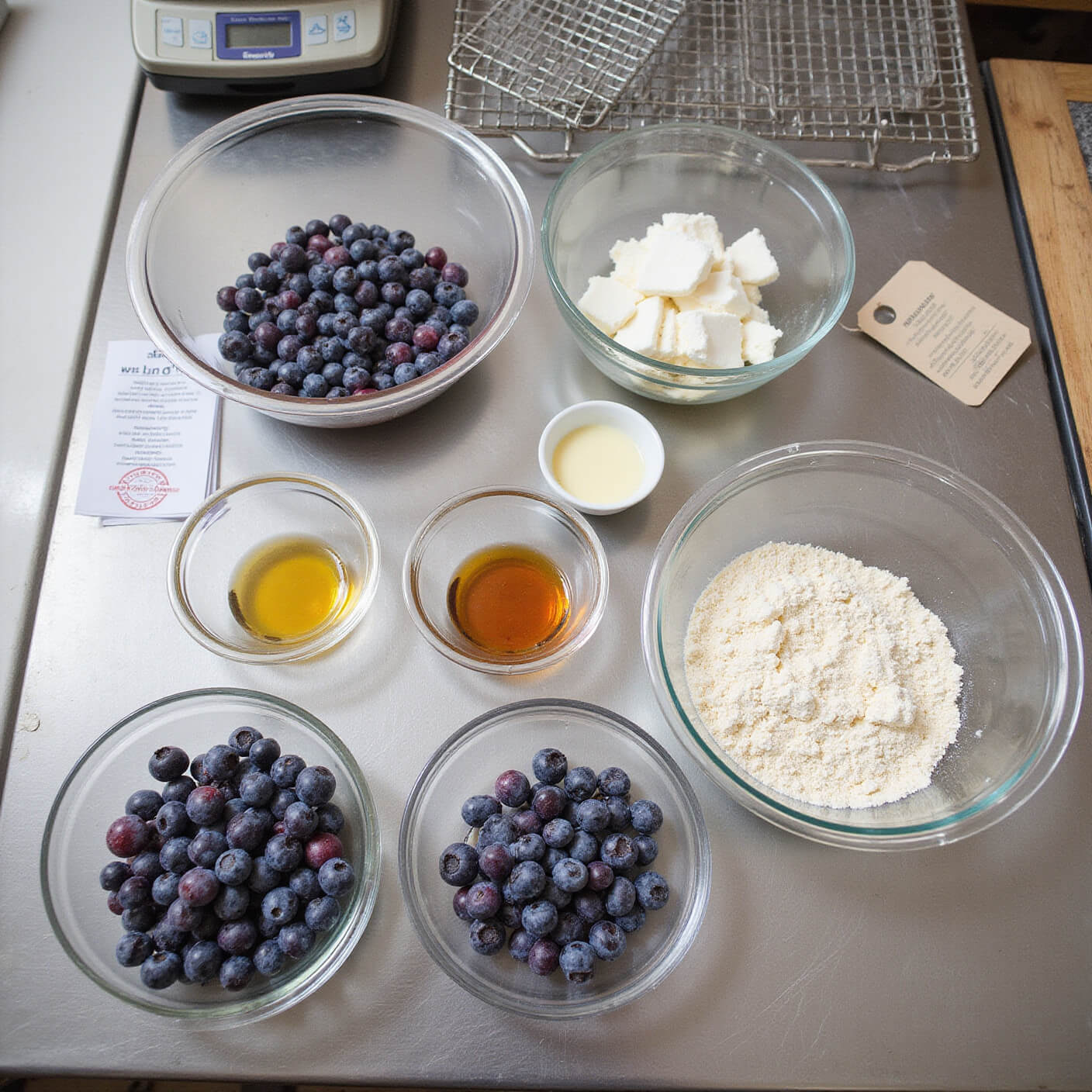 Overhead view of a professional kitchen prep station with bowls of blueberries, almond flour, honey, and coconut oil, organized on a stainless steel surface with a kitchen scale, recipe cards, and cooling racks visible.