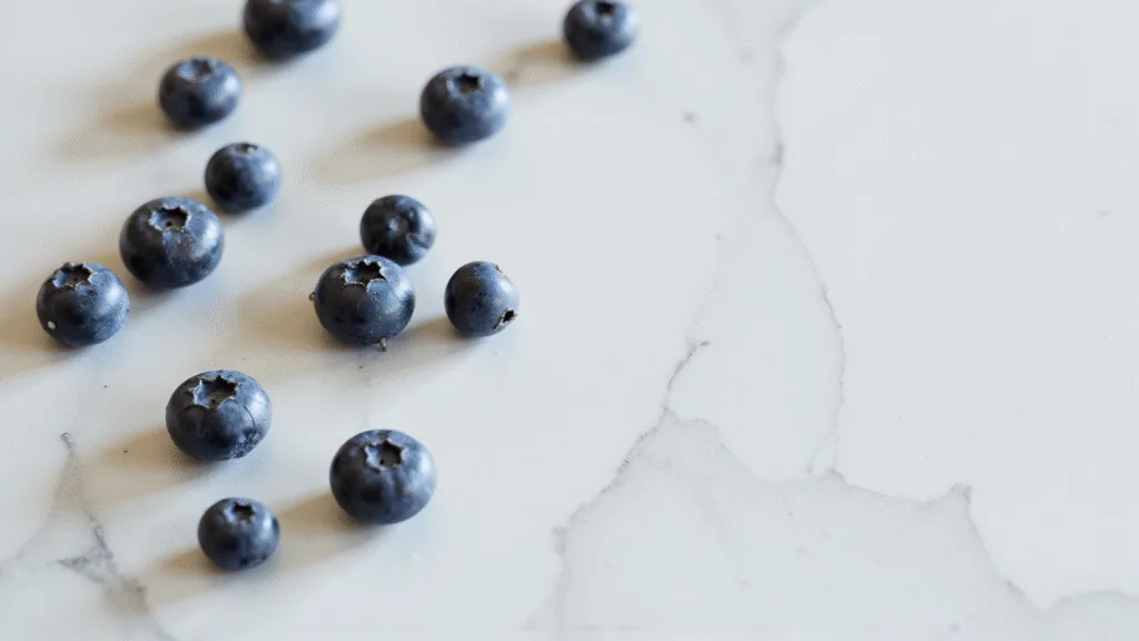 "Fresh blueberries scattered on a white marble surface with morning sunlight casting soft shadows and glistening dew drops."